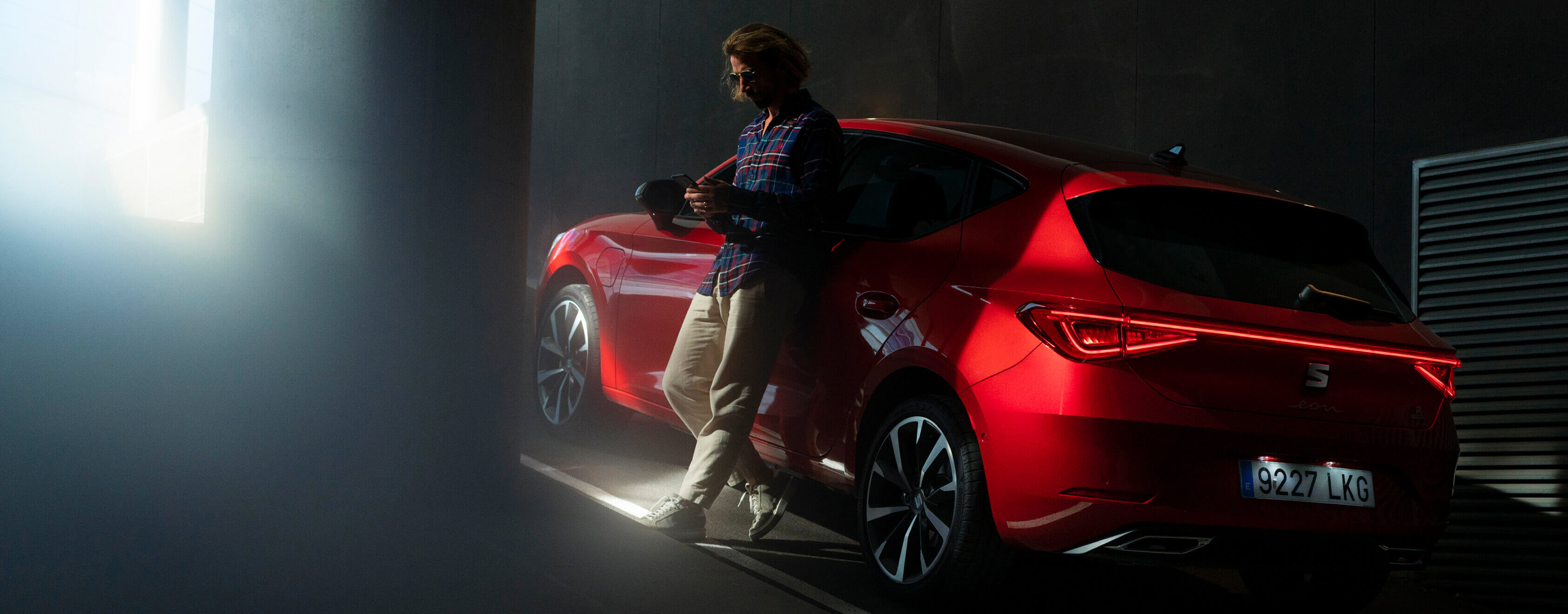 Red SEAT Leon parked in an indoor parking area. A man leans against the driver's side door, using a mobile device.